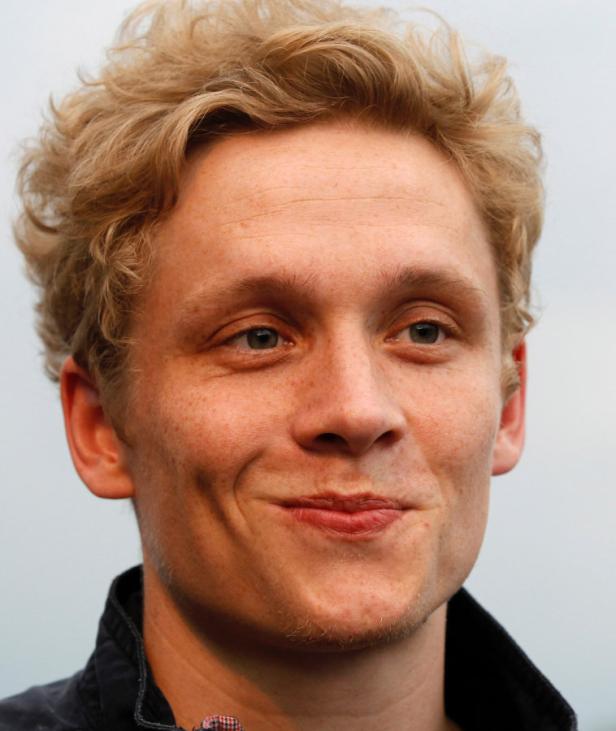 Ein Mann mit blonden, lockigen Haaren lächelt leicht.