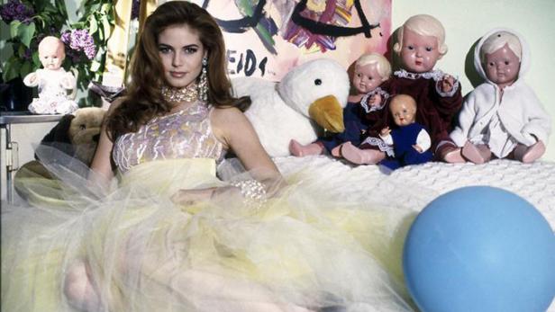 Eine Frau in einem gelben Kleid posiert mit Puppen und einem Stofftier auf einem Bett.