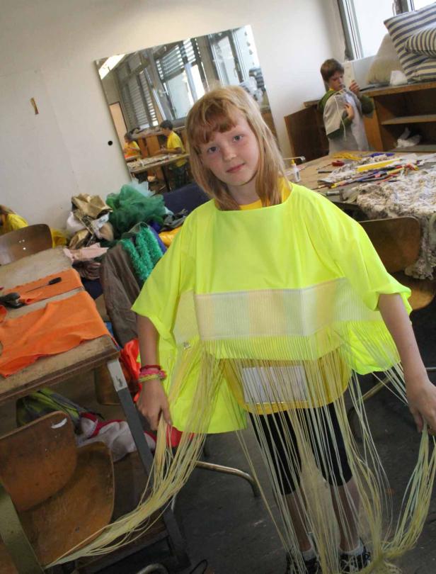 Ein Mädchen trägt ein neonfarbenes Kleid mit Fransen in einem chaotischen Raum.