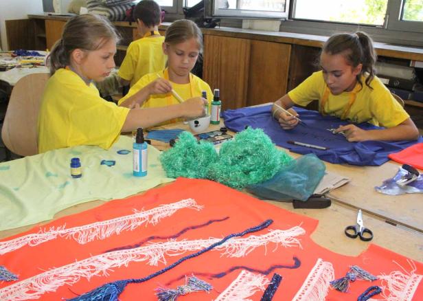 Drei Mädchen bemalen Stoffe in verschiedenen Farben mit Pinseln.