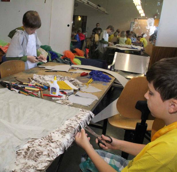 Kinder sitzen an Tischen und basteln mit Stoff, Scheren und Klebstoff in einem Werkraum.