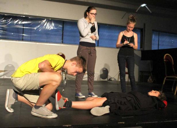 Auf einer Bühne untersucht ein junger Mann mit Stethoskop eine am Boden liegende Person, während zwei Frauen zusehen.