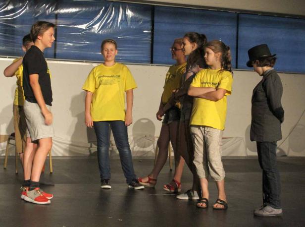 Eine Gruppe von Kindern steht auf einer Bühne, einige tragen gelbe T-Shirts mit der Aufschrift „KinderuniKunst“.