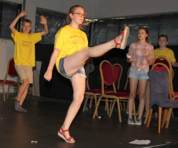 Ein junges Mädchen tritt auf einer Bühne in die Luft, während andere Jugendliche zusehen.