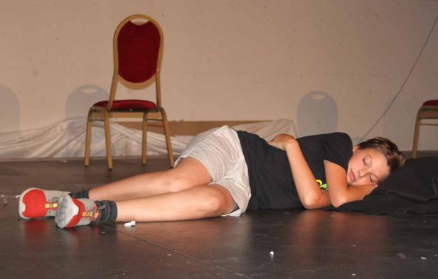 Ein Junge schläft auf einer Bühne, neben ihm steht ein roter Stuhl.