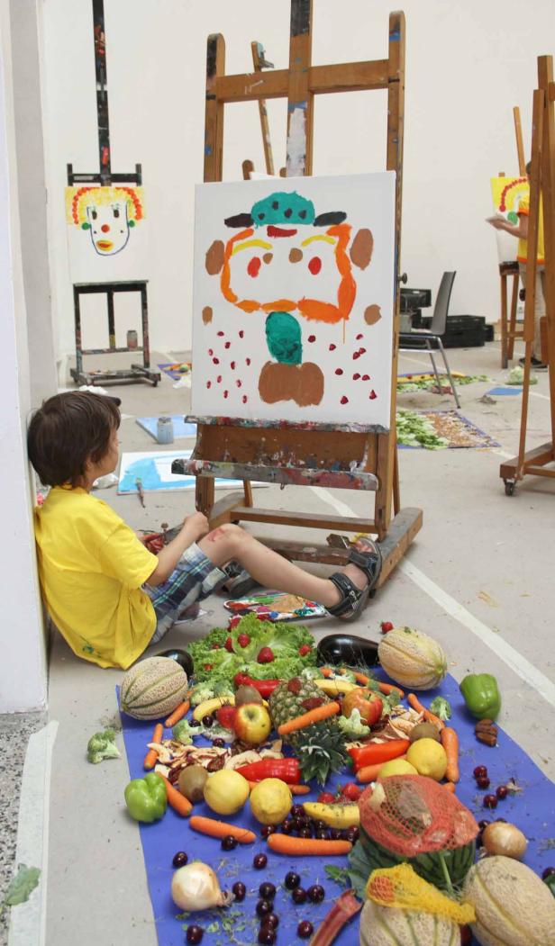 Ein Junge sitzt neben einer Leinwand mit einem bunten, abstrakten Gesicht, umgeben von Obst und Gemüse.