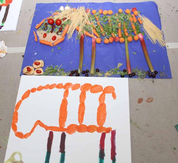 Ein Stillleben mit Gemüse, Obst und Nudeln auf einem blauen Tuch und ein gemaltes Bild darunter.