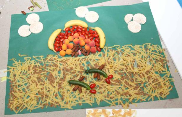 Ein grünes Papierbild zeigt eine Landschaft aus Bananen, Tomaten, Aprikosen, Datteln, Reiswaffeln, Nudeln und Gurken.