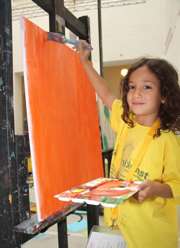 Ein junges Mädchen malt mit einem Pinsel ein orangefarbenes Bild auf einer Staffelei.