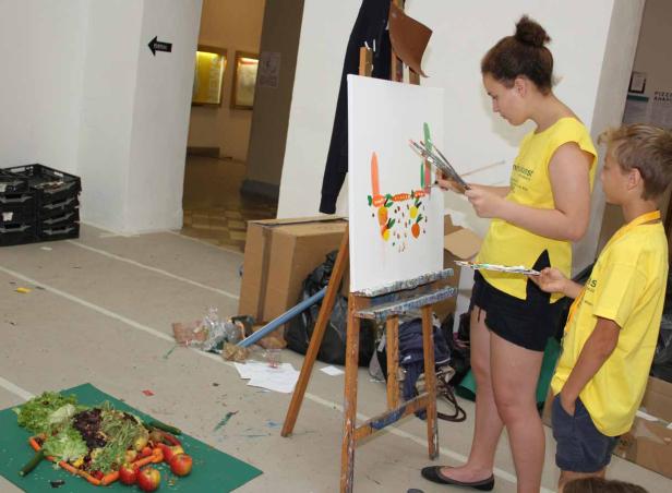 Ein Mädchen und ein Junge malen ein Stillleben mit Obst und Gemüse auf eine Leinwand.