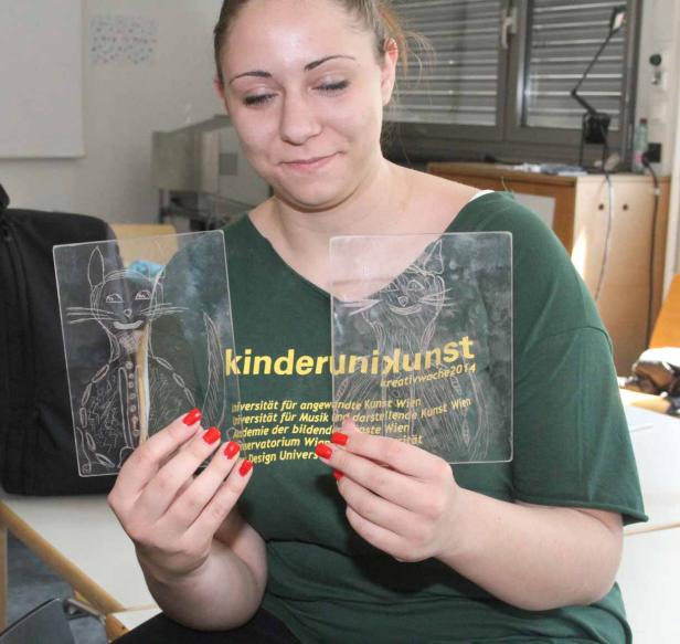 Eine Frau hält zwei Acrylglasplatten mit Katzenzeichnungen und dem Schriftzug „kinderuniKunst“.