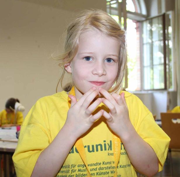 Ein blondes Mädchen mit blauem Blick trägt ein gelbes T-Shirt und hält die Hände vor dem Mund.