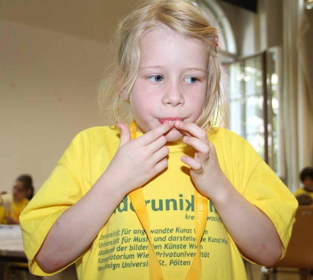 Ein blondes Mädchen im gelben T-Shirt mit dem Finger im Mund.