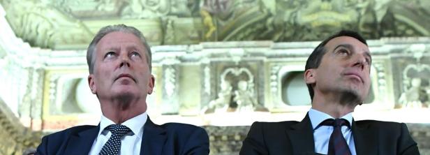 Zwei Männer in Anzügen blicken nach oben, im Hintergrund ein verziertes Deckengemälde.