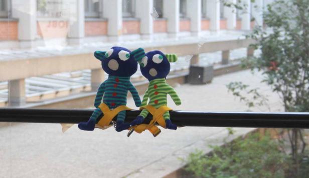 Zwei Stofftiere mit Gurten sitzen auf einer Stange vor einem Fenster.