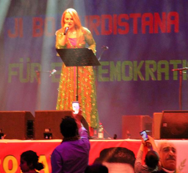 Eine Frau singt auf einer Bühne vor einem Banner mit der Aufschrift „Für Kurdistan, für Demokratie“.