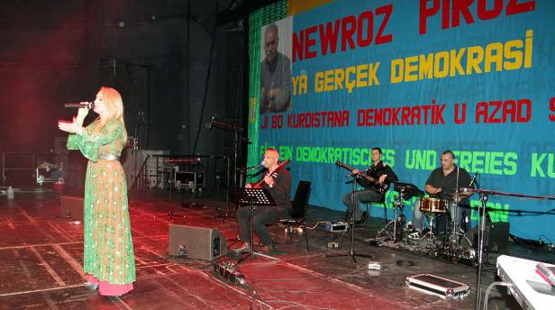 Eine Sängerin tritt mit ihrer Band vor einem Banner mit kurdischen Aufschriften auf einer Bühne auf.