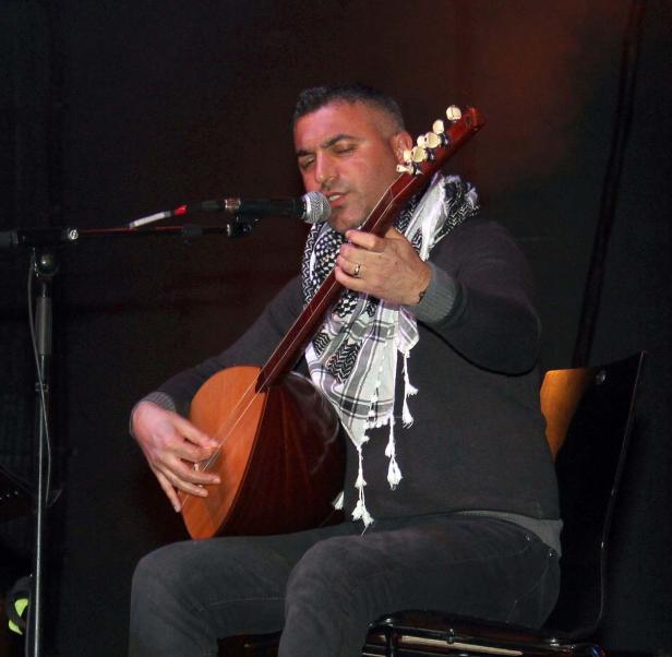 Ein Mann mit Schal sitzt und spielt ein Saiteninstrument vor einem Mikrofon.