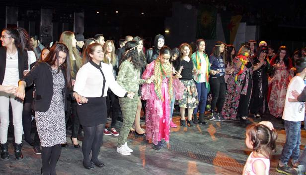 Eine Gruppe von Menschen hält Hände und tanzt auf einer Bühne, im Hintergrund eine kurdische Flagge.