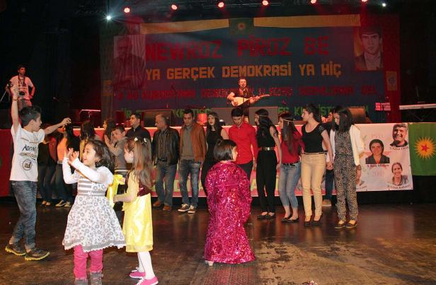 Kinder tanzen auf einer Bühne vor einer kurdischen Flagge und einem Transparent mit der Aufschrift „Newroz Pîroz Be“.