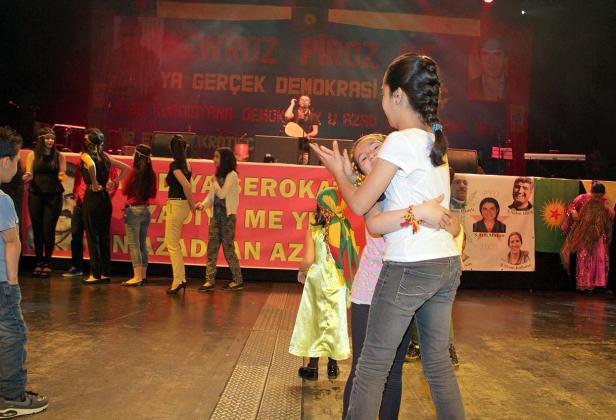 Kinder tanzen auf einer Bühne vor einem Banner mit kurdischen Schriftzeichen.