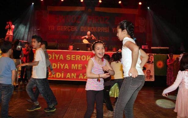 Kinder tanzen auf einer Bühne vor einem Banner mit kurdischen Aufschriften.
