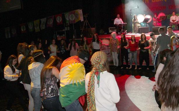 Eine Gruppe von Menschen tanzt auf einer Bühne mit kurdischen Flaggen im Hintergrund.