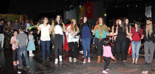 Eine Gruppe von Menschen, darunter Kinder, tanzt gemeinsam vor kurdischen Flaggen.