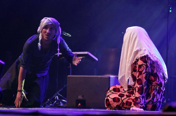 Ein Mann mit Turban gestikuliert neben einer knienden, verschleierten Frau auf einer Bühne.