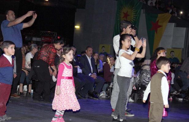Eine Gruppe von Menschen, darunter Kinder, bei einer Veranstaltung mit kurdischen Flaggen im Hintergrund.