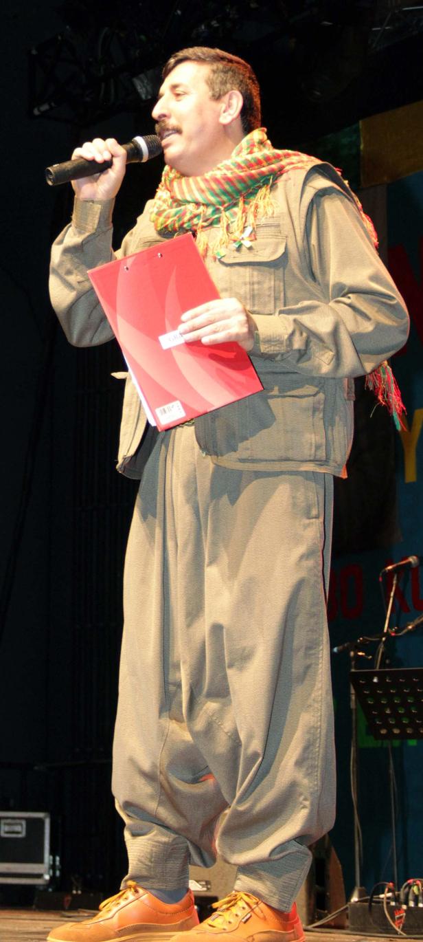 Ein Mann mit Schnurrbart hält ein Mikrofon und eine rote Mappe auf einer Bühne.