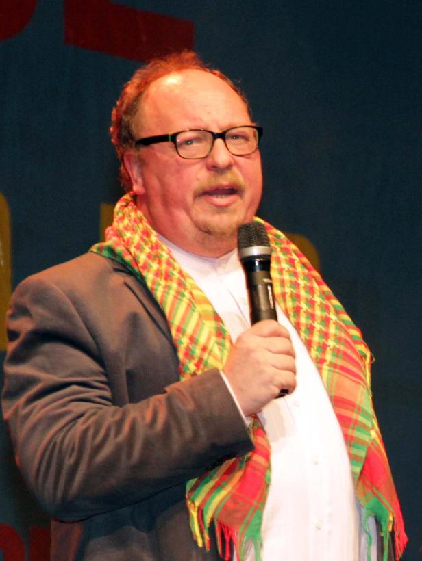 Ein Mann mit Brille und kariertem Schal hält ein Mikrofon in der Hand.