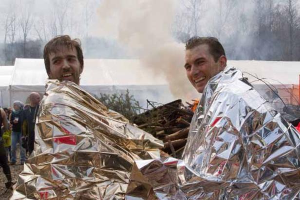 Zwei Männer, in Rettungsdecken gehüllt, stehen vor einem Lagerfeuer.