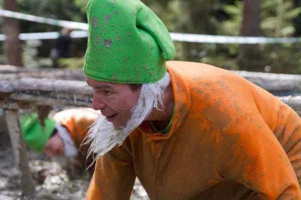 Ein Mann mit grünem Hut und Bart kriecht unter einem Hindernis bei einem Rennen.