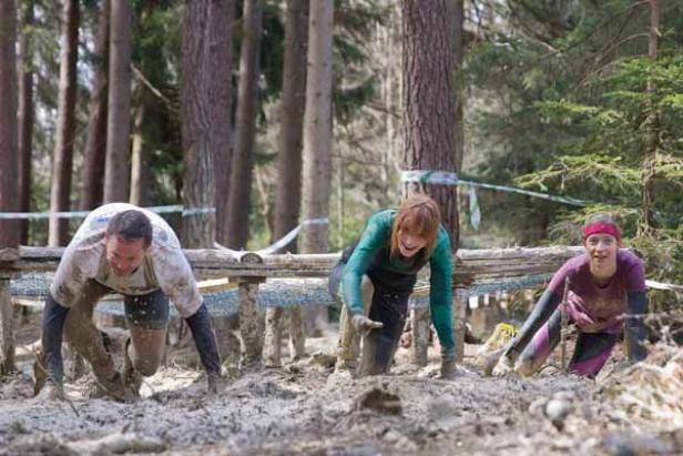 Drei Personen kriechen unter einem Hindernis bei einem Schlammlauf hindurch.