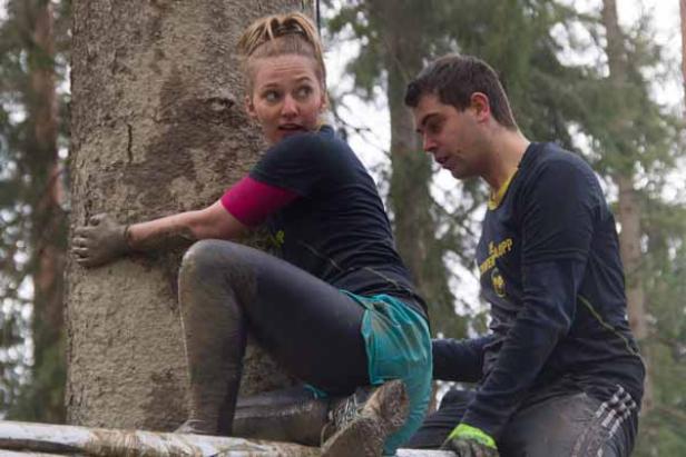 Eine Frau klettert mit schlammverschmierten Händen einen Baum hoch, während ein Mann ihr hilft.