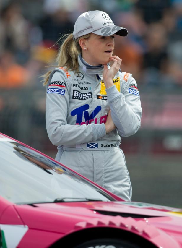 Susie Wolff in Rennfahrerkluft steht nachdenklich an einem pinkfarbenen Rennwagen.