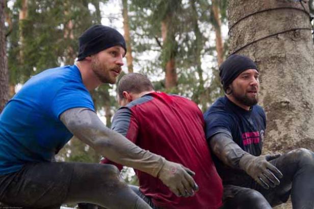 Drei Männer mit Mützen und schmutziger Kleidung sitzen im Wald.