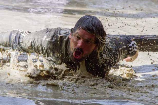 Ein Mann kriecht unter einem Hindernis in einem schlammigen Wasserbecken hindurch.