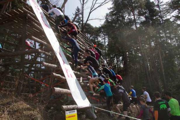 Teilnehmer klettern bei einem Hindernislauf eine steile Holzkonstruktion hoch.