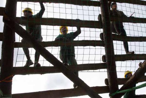 Mehrere Bauarbeiter mit Schutzhelmen klettern auf einem Holzgerüst mit Netz.
