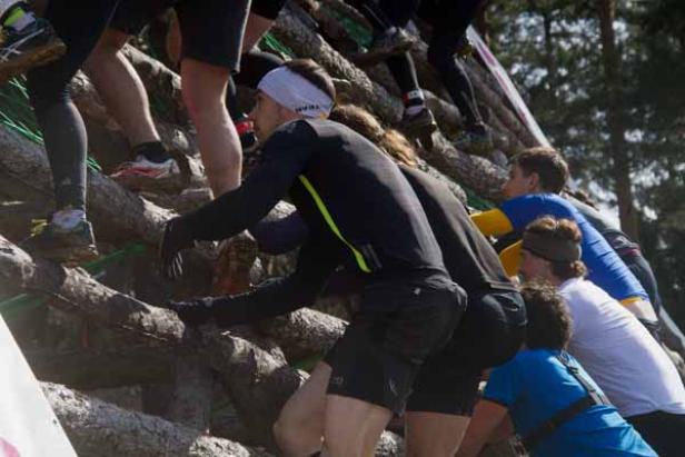 Teilnehmer klettern bei einem Hindernislauf über eine hohe Holzwand.