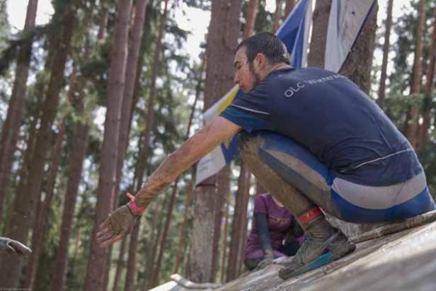 Ein schlammverschmierter Mann klettert in einem Wald über einen Baumstamm.