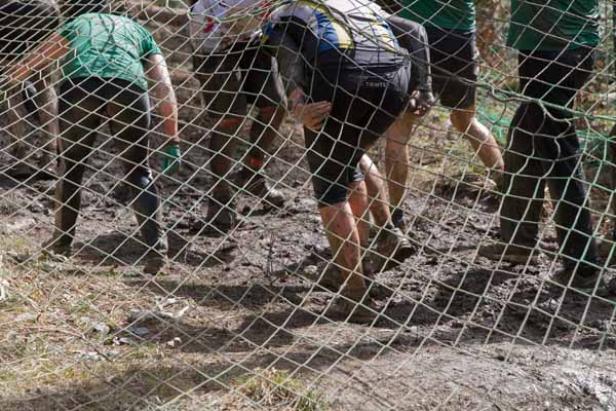 Mehrere Personen kriechen unter einem Netz in schlammigem Gelände hindurch.