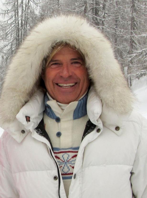 Ein lächelnder Mann mit weißer Winterjacke und Fellkapuze vor verschneiten Bäumen.