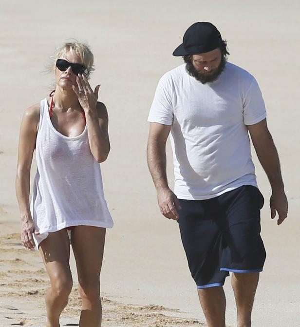 Pamela Anderson und ein Mann mit Bart spazieren am Strand entlang.