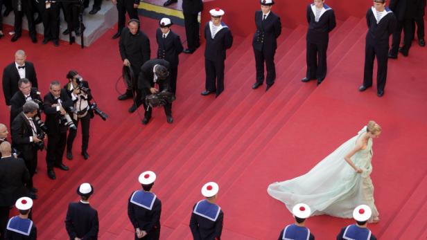 Eine Frau in einem hellen Kleid geht eine rote Treppe hinunter, umgeben von Fotografen und Marineuniformierten.