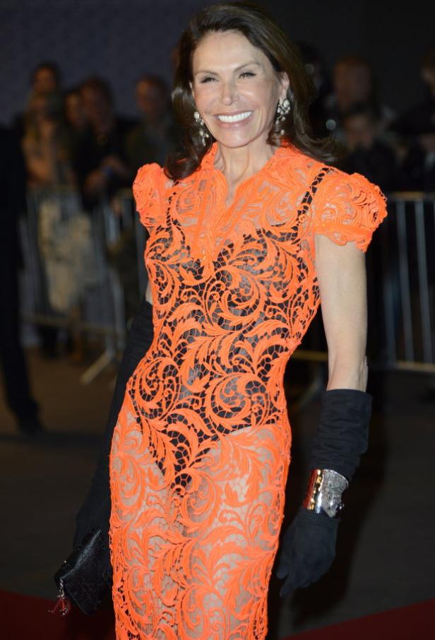 Eine Frau in einem orangefarbenen Spitzenkleid und schwarzen Handschuhen lächelt.