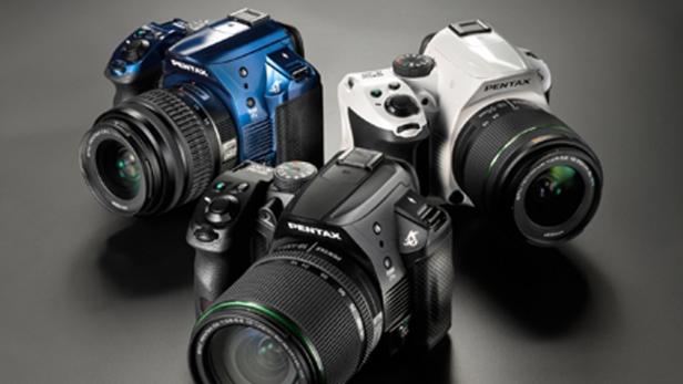 Drei Pentax-Kameras in den Farben Schwarz, Blau und Weiß liegen auf einem Tisch.