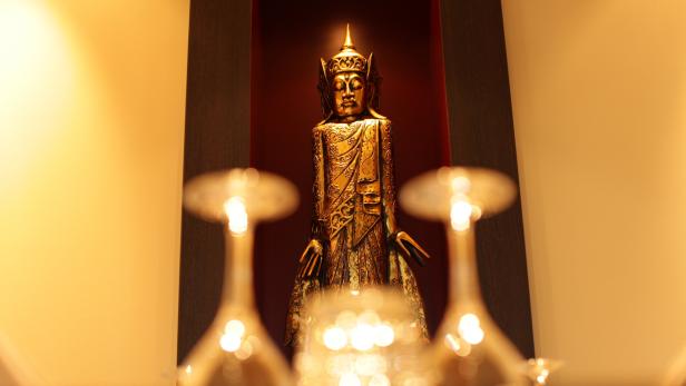 Eine goldene Buddha-Statue steht in einer Nische, teilweise verdeckt durch Weingläser.